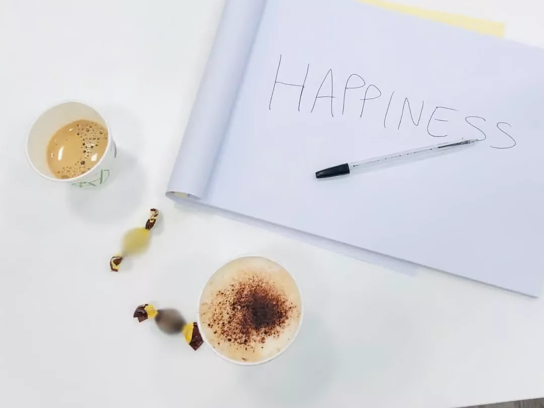 Un cappuccino e un caffè al ginseng da VICINOtea, serviti con due cioccolatini, vicino a un taccuino su cui campeggia la parola "Happiness"